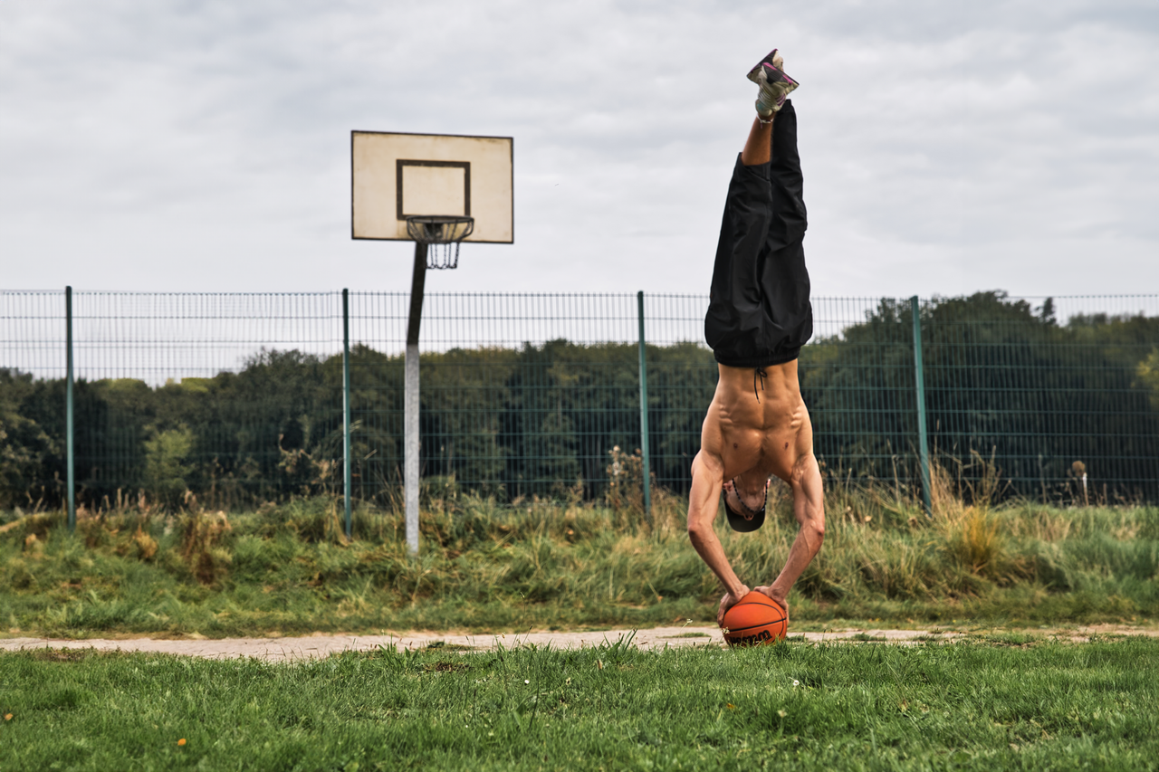 Athletic man doing a handstand with a basketball on an outdoor court, showcasing strength and balance.